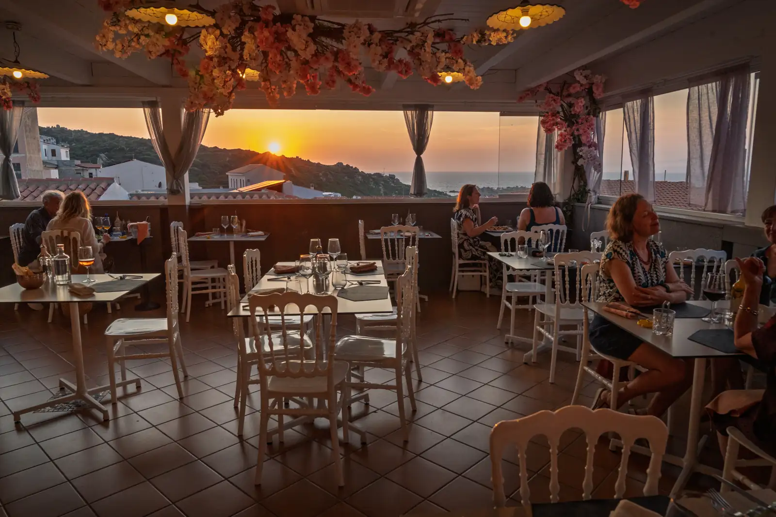Die Tische der Terrasse bei Sonnenuntergang, mit sitzenden Gästen und der Sonne über dem Meer